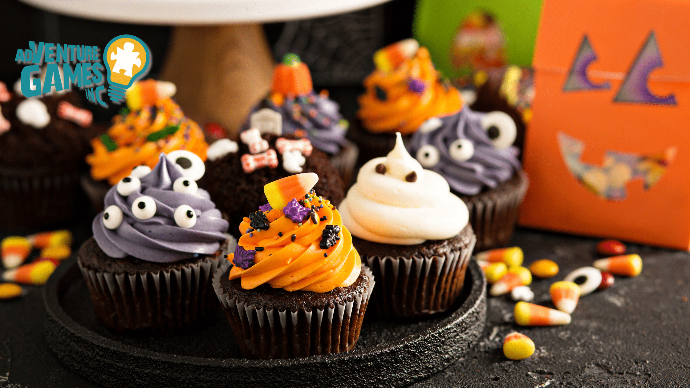 Close-up of Halloween-themed cupcakes with orange, purple, and white frosting decorated with candy eyes, candy corn, and spooky sprinkles, displayed on a dark tray with the Adventure Games Inc. logo.
