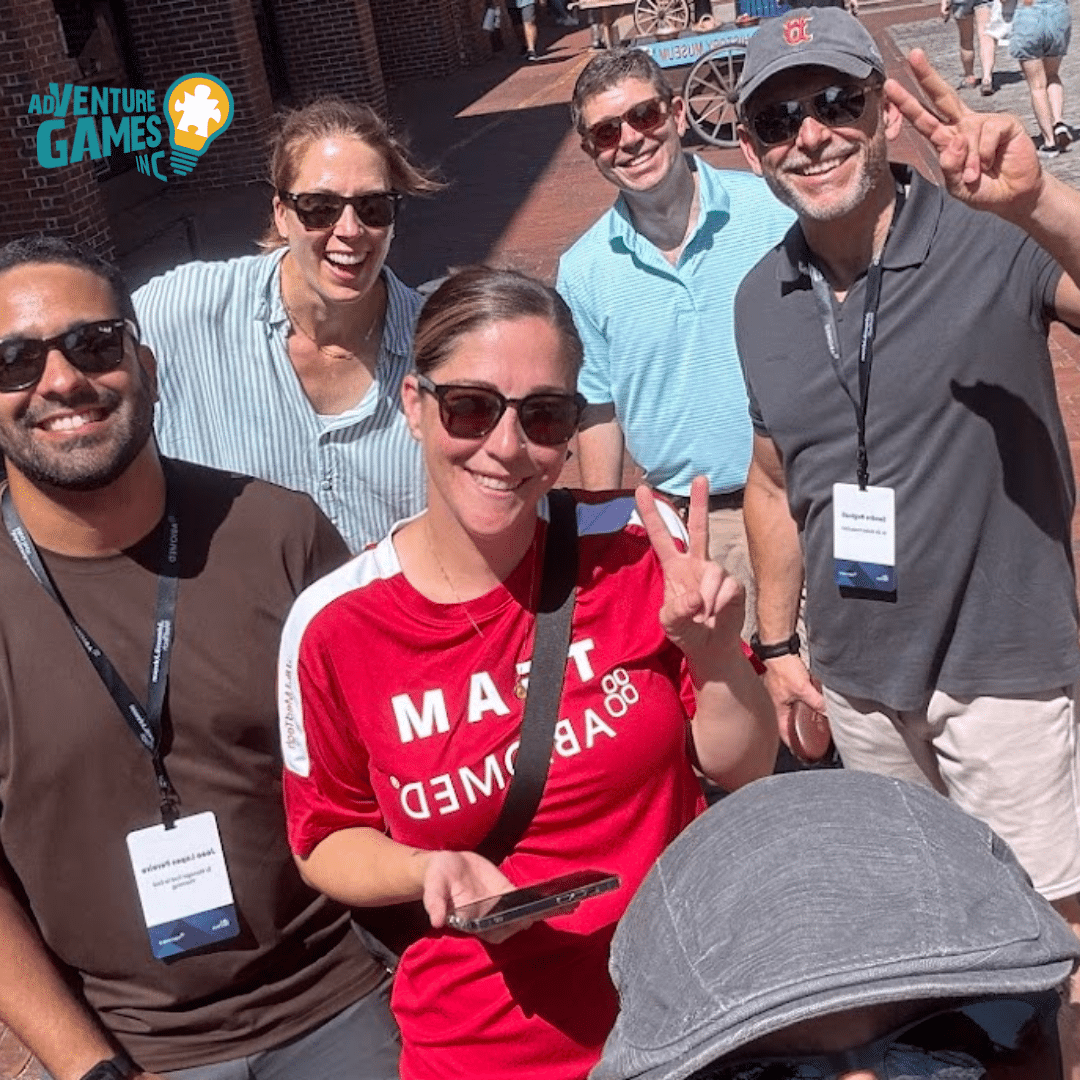 A smiling group of six people pose for a selfie during a sunny outdoor team-building activity. They are wearing lanyards and casual clothes, with one person in a red “TEAM” shirt making a peace sign. Brick buildings and cobblestone streets are visible in the background, with the Adventure Games Inc. logo in the top left corner.
