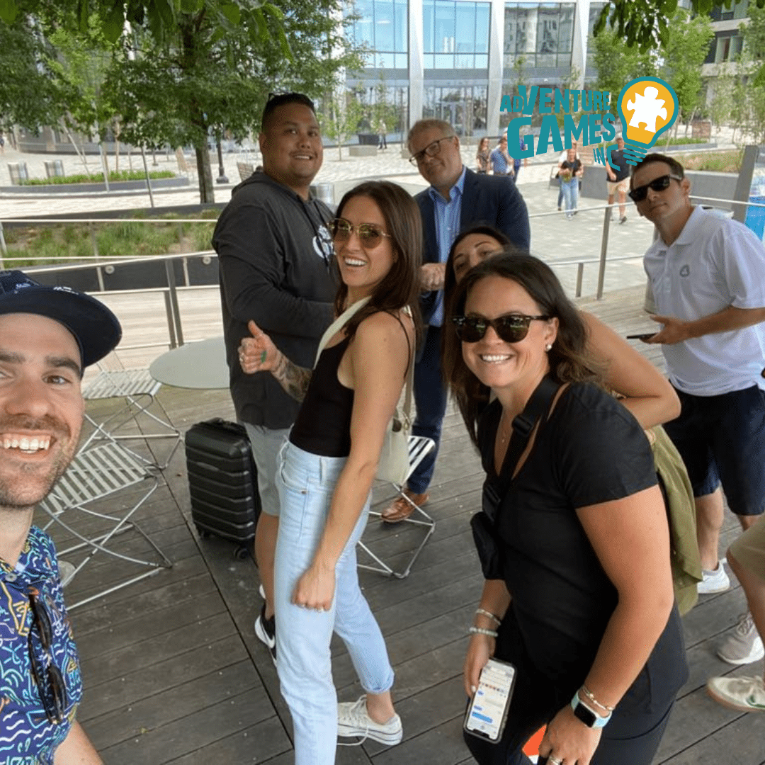 A group of seven coworkers smile and pose together outdoors on a wooden patio surrounded by modern buildings and trees. They appear to be participating in a team-building activity, with some giving thumbs-up and holding phones or small bags. The Adventure Games Inc. logo is displayed in the upper right corner.