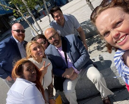A group of seven coworkers pose for a cheerful outdoor selfie on a sunny day in the city. They are smiling and gathered closely together, some standing and some sitting on a stone bench. The group appears to be taking part in a team-building activity, with buildings, trees, and a sidewalk visible in the background.