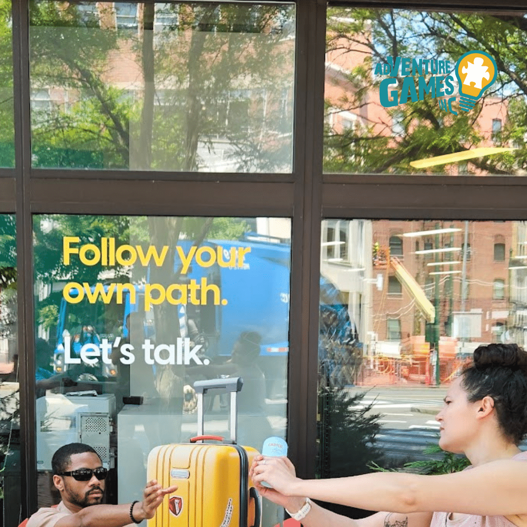 Two participants engage in an outdoor team-building challenge in front of a glass building with the words “Follow your own path. Let’s talk.” printed on the window. One person holds a yellow suitcase while another reaches out toward them. The Adventure Games Inc. logo is displayed in the upper right corner.