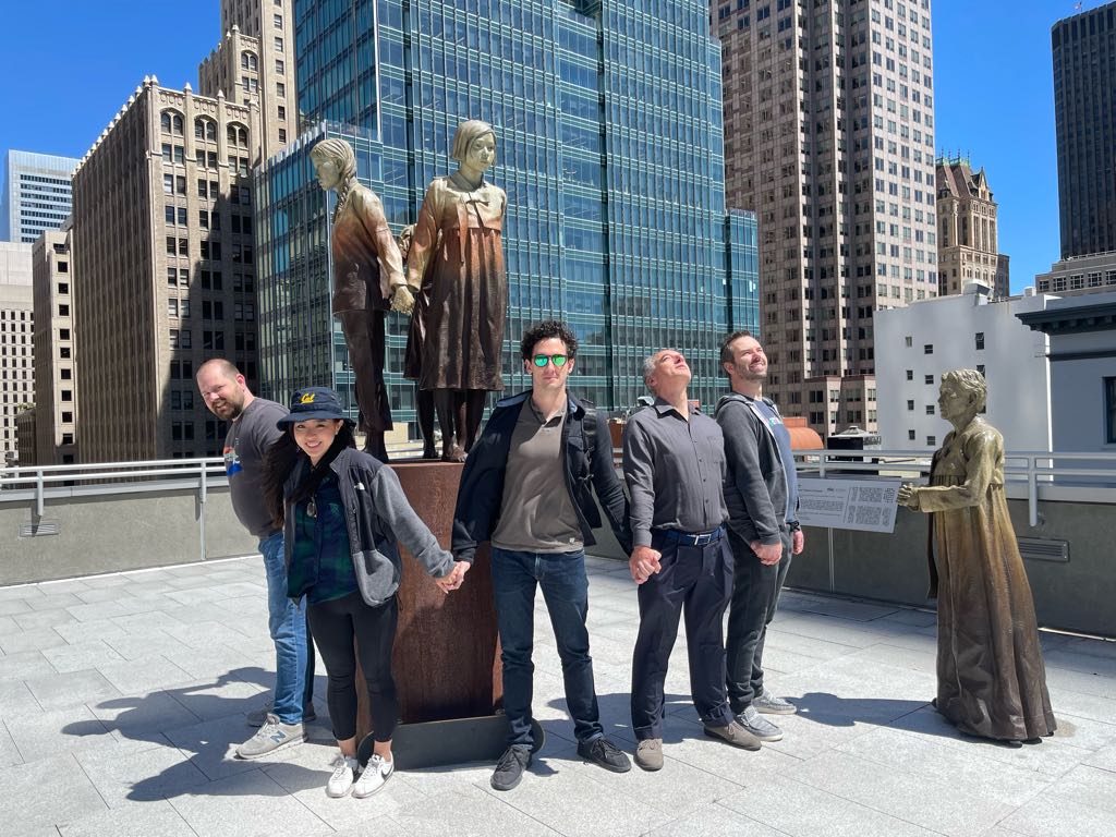 A team poses hand-in-hand with bronze statues on a rooftop in San Francisco’s Financial District during a creative team building challenge led by Adventure Games Inc, the best team building company in San Francisco.