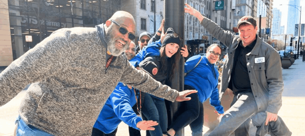 Group of coworkers in bright blue and gray jackets posing mid-action on a city sidewalk during a high-energy team-building scavenger hunt by Adventure Games Inc.