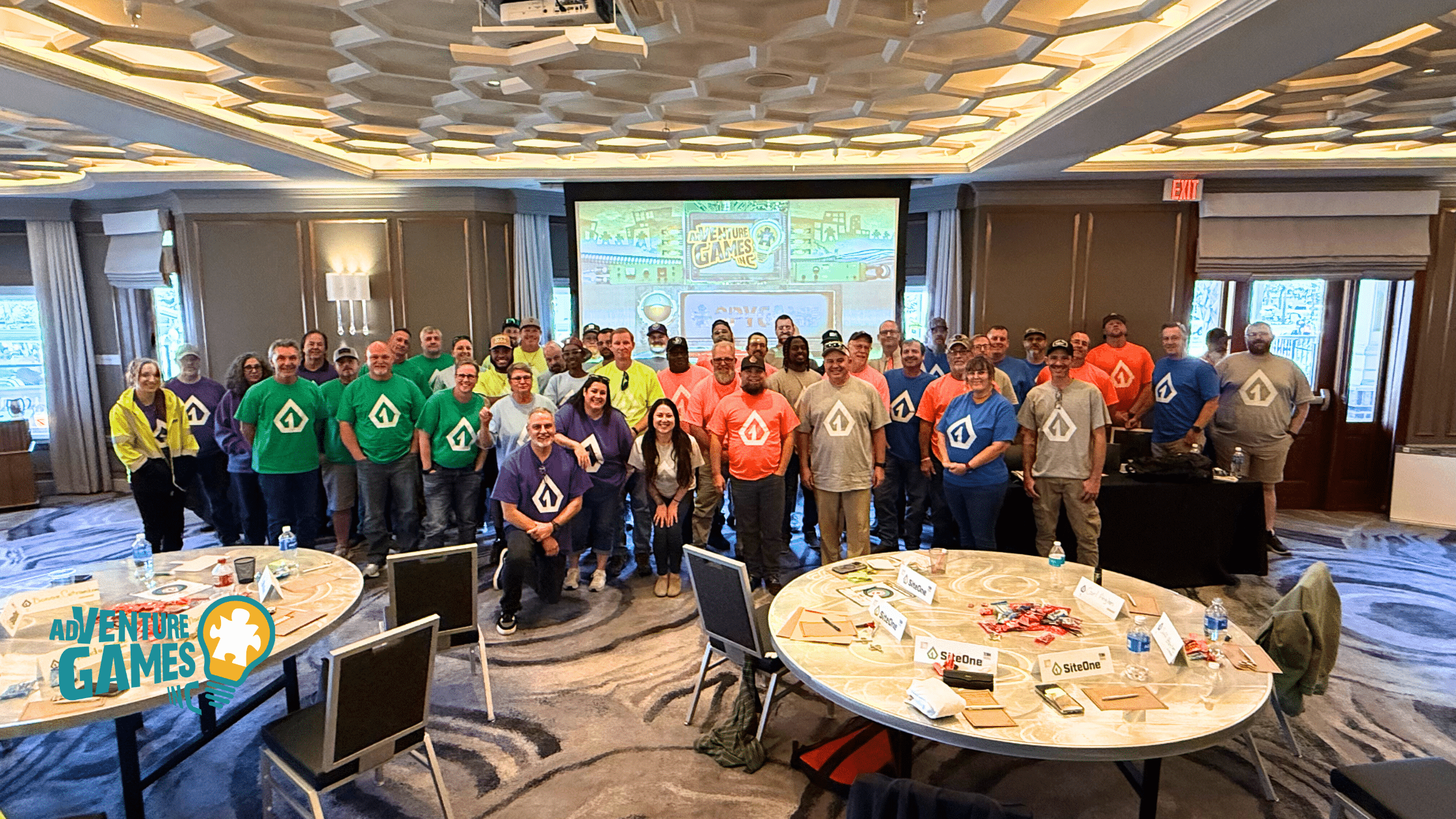 Large team of corporate employees wearing color-coded shirts after completing a group team-building activity led by Adventure Games Inc., posing in a hotel conference room.