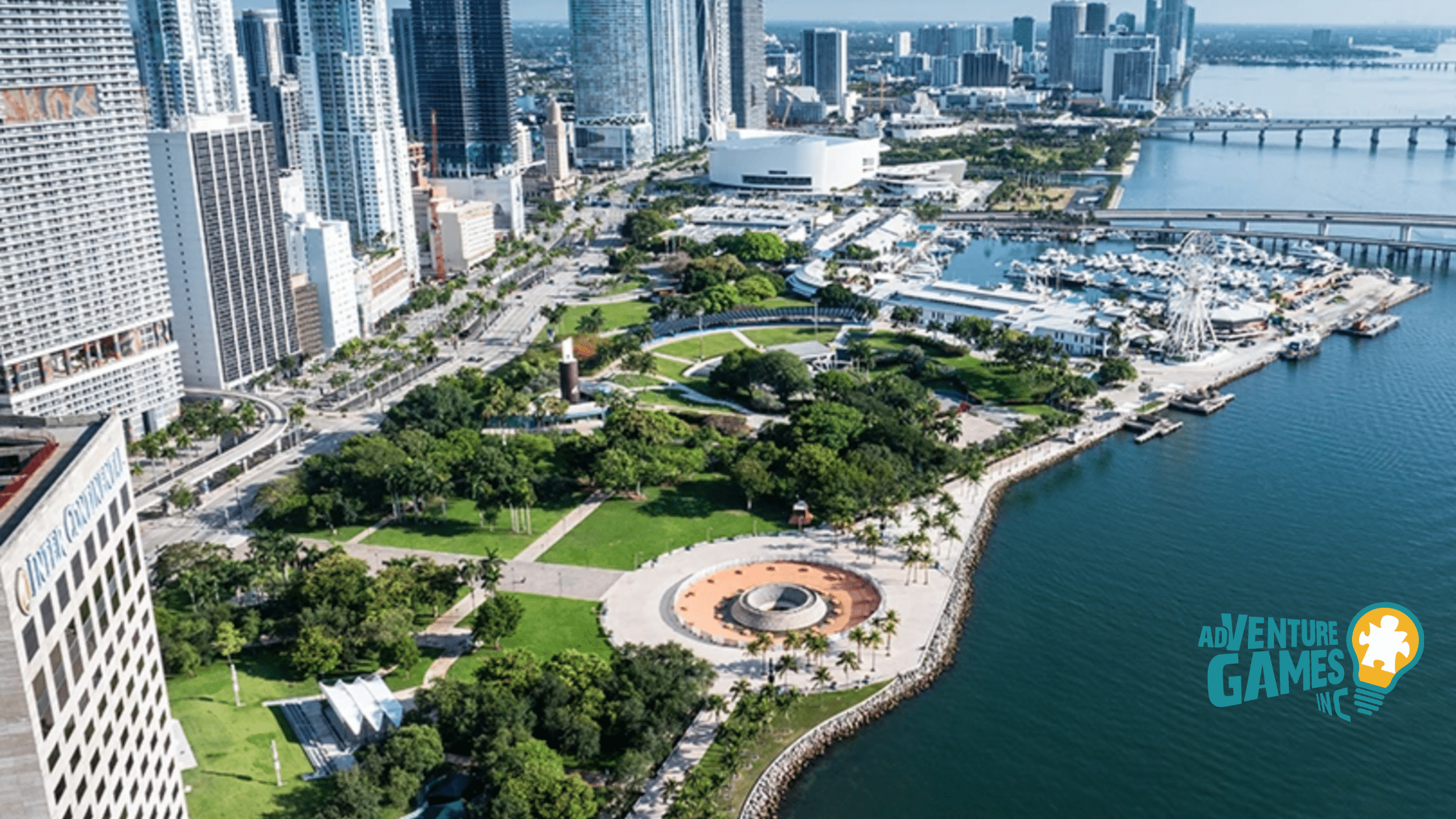 Aerial view of Bayfront Park in Miami Florida, showing the circular Bandshell plaza, shaded lawns, waterfront promenade along Biscayne Bay and the downtown skyline in the background.
