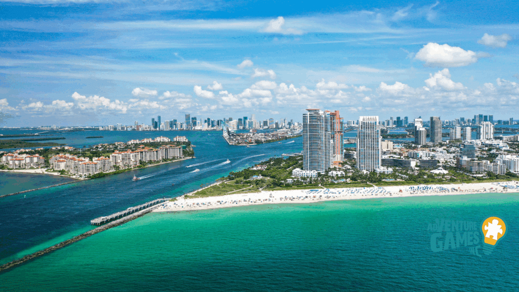 Aerial view of South Pointe Park in Miami Beach Florida, showing the sandy shoreline, green park lawns, a long pier extending into turquoise water, and the Miami skyline on the horizon.