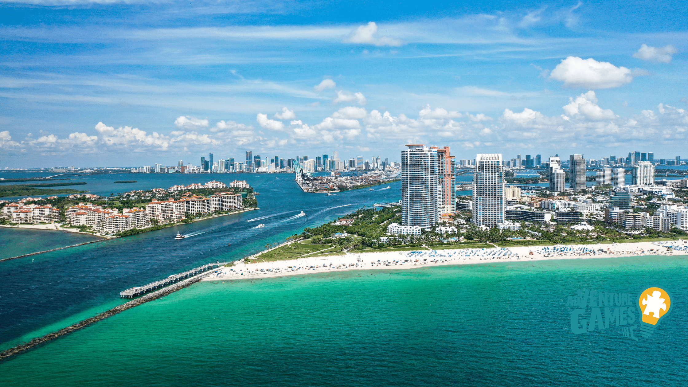 Aerial view of South Pointe Park in Miami Beach Florida, showing the sandy shoreline, green park lawns, a long pier extending into turquoise water, and the Miami skyline on the horizon.