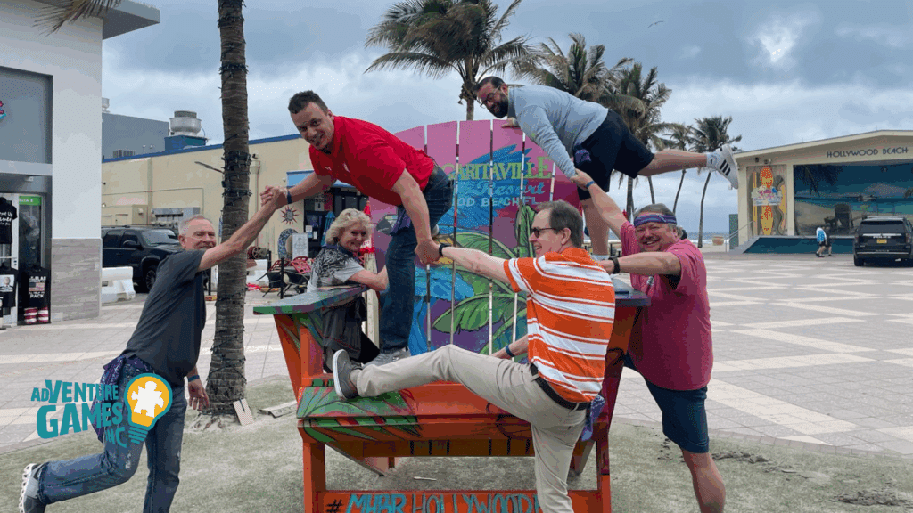 A corporate team balances and laughs while posing on a giant beach chair during an outdoor team building activity hosted by Adventure Games Inc. in Hollywood Beach, Florida.