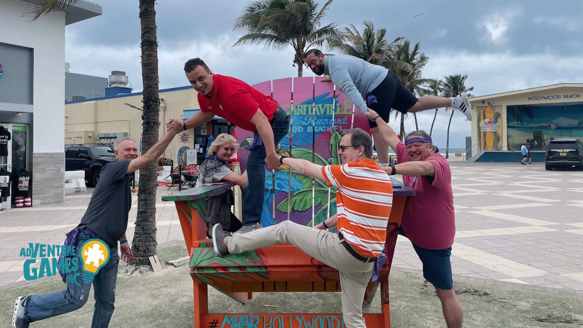A corporate team balances and laughs while posing on a giant beach chair during an outdoor team building activity hosted by Adventure Games Inc. in Hollywood Beach, Florida.