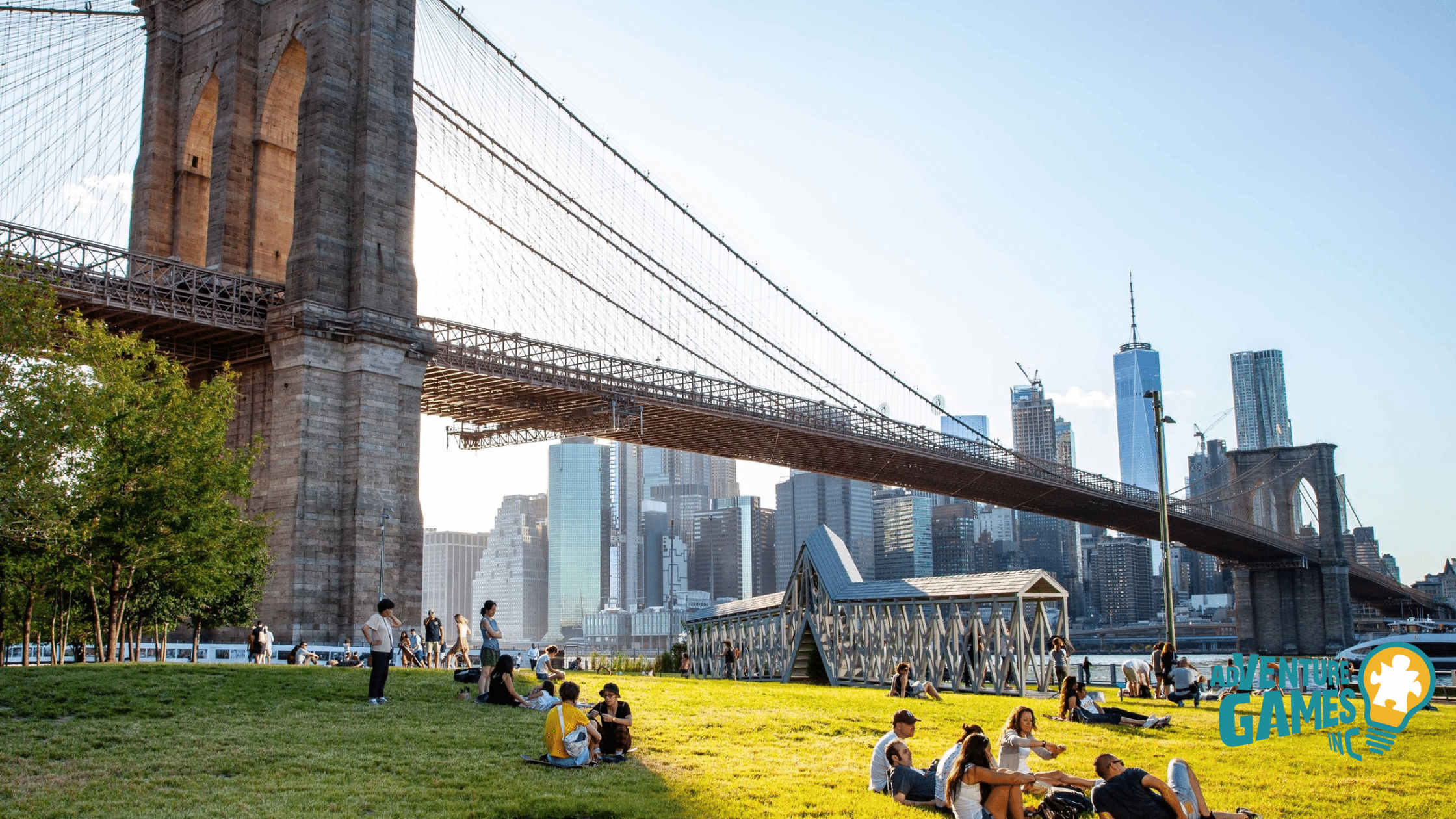 People relaxing on the lawn beneath the Brooklyn Bridge with the Lower Manhattan skyline in view.