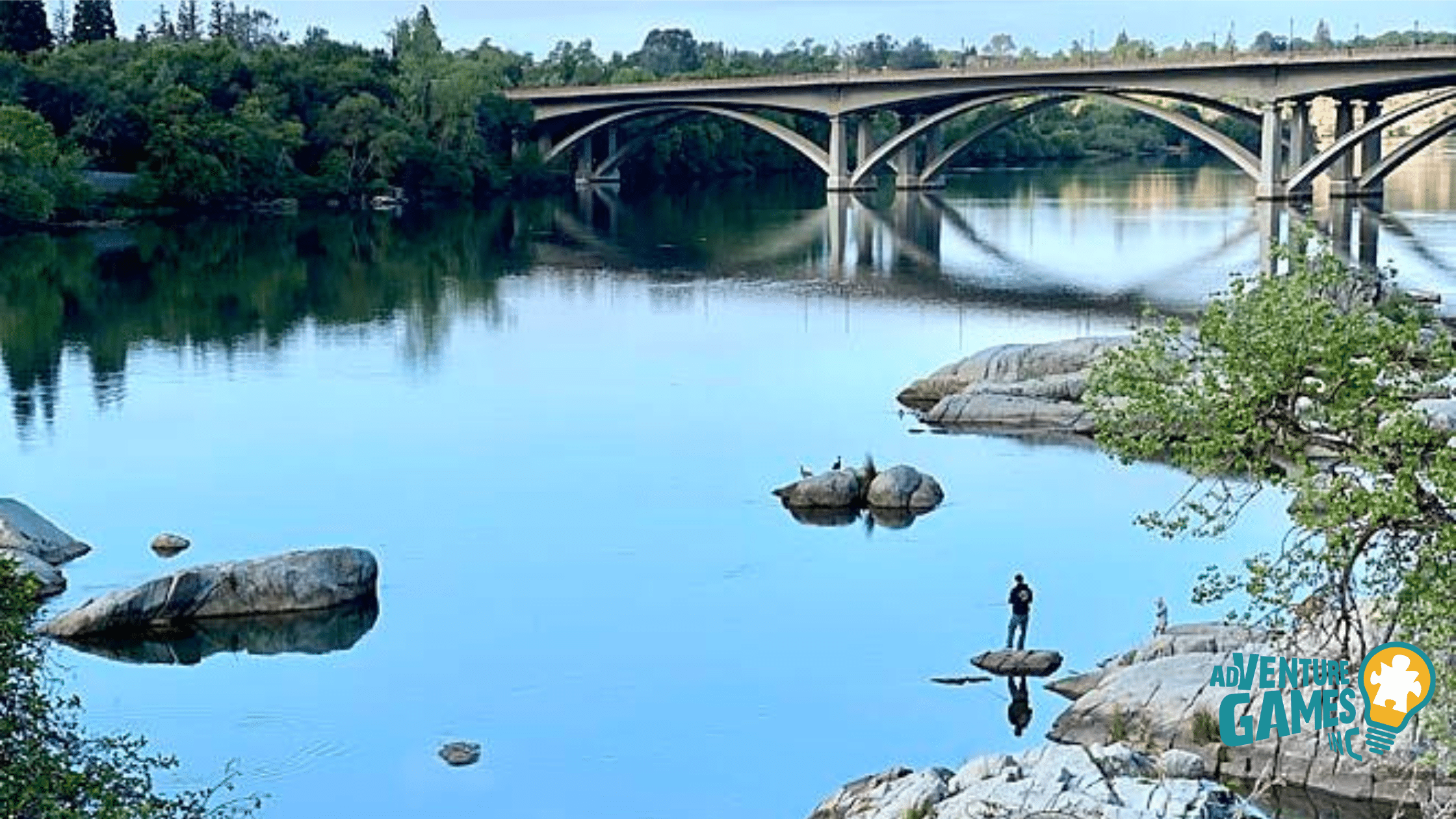 Calm river with granite boulders and an arched bridge reflected in the water along Sacramento’s American River Parkway.