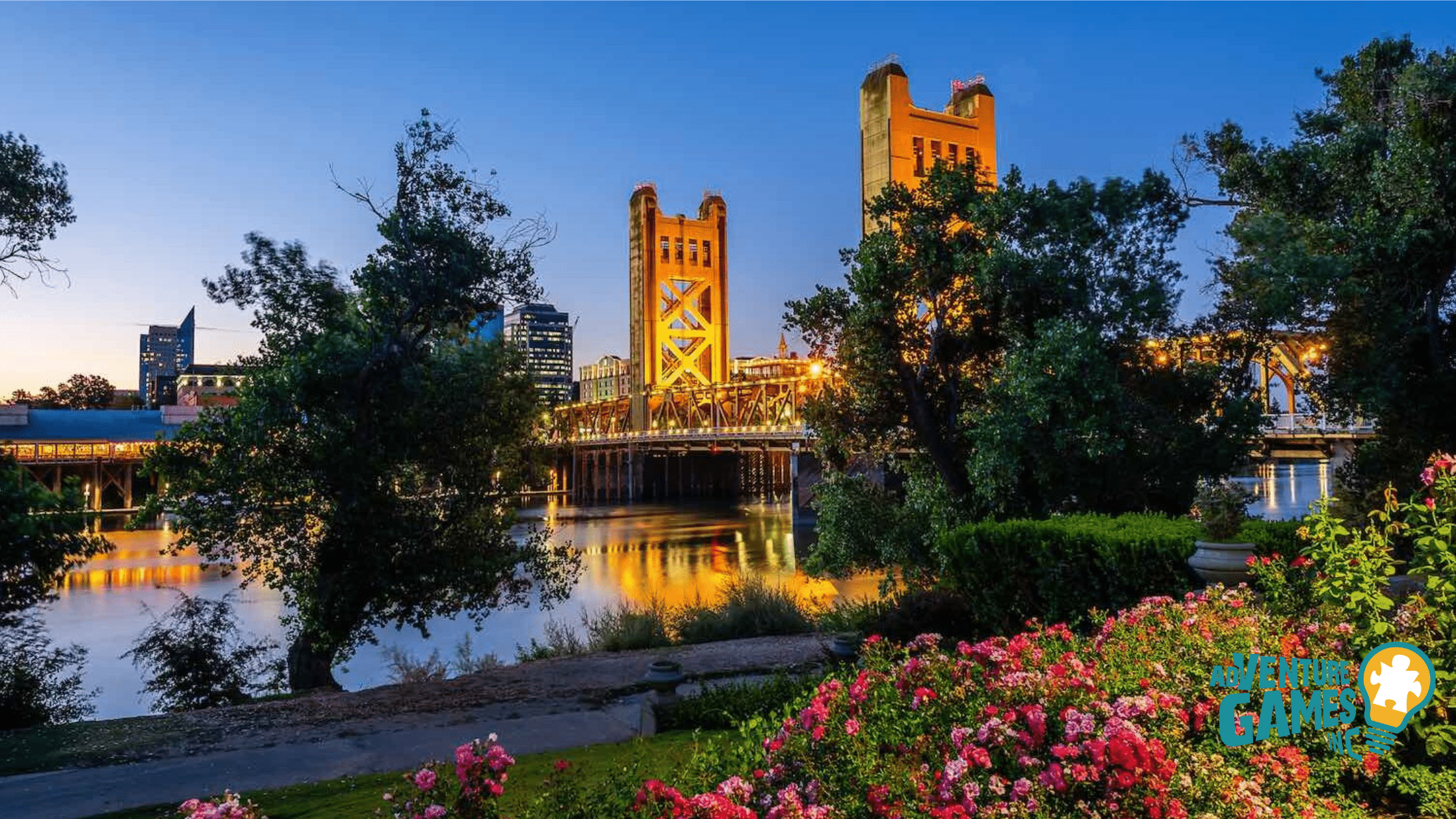 Tower Bridge glowing at dusk over the Sacramento River, viewed from a flowered riverfront path.