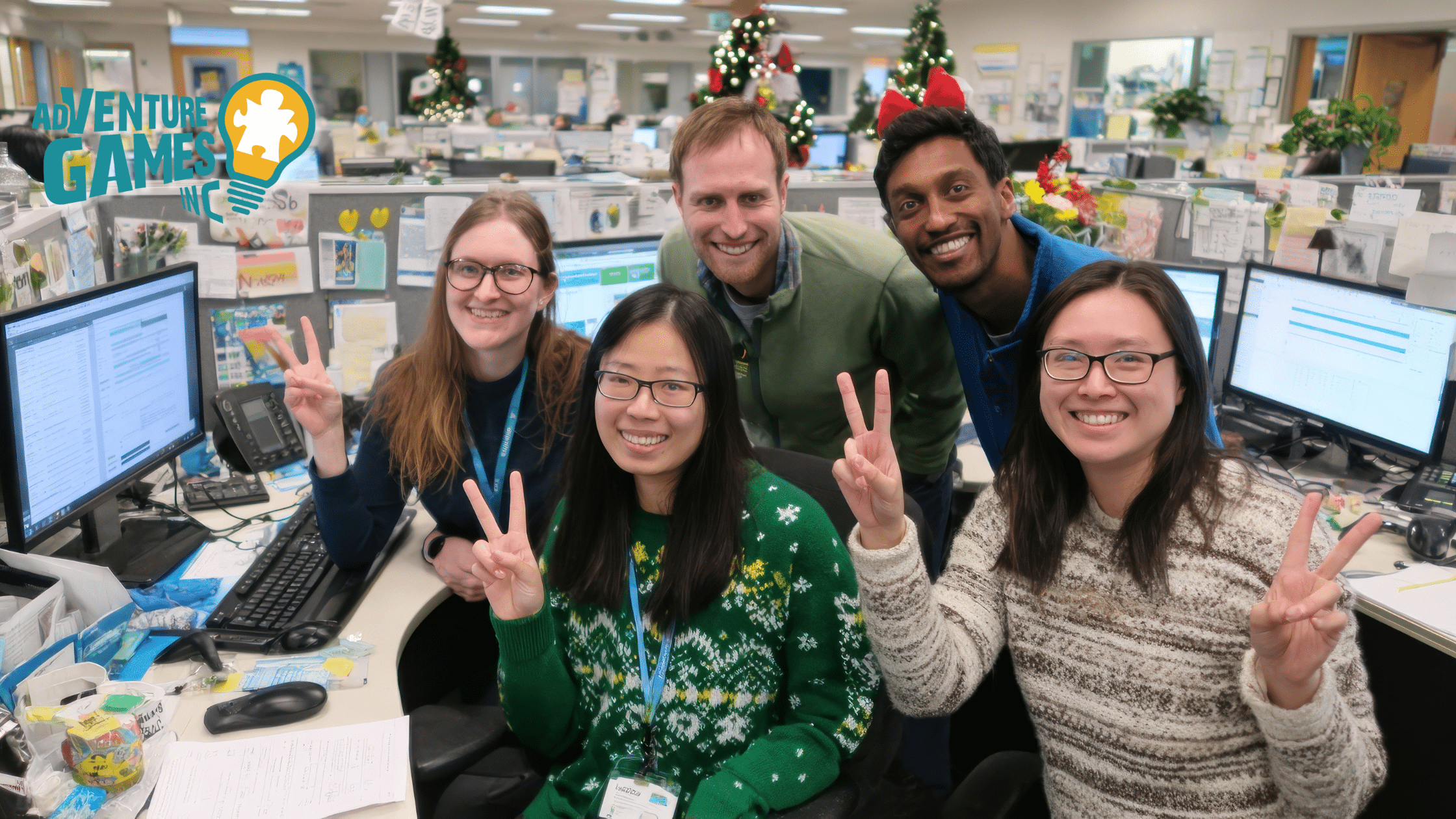 A group of five coworkers smiling and holding up peace signs in a decorated office with Christmas trees in the background, sitting at desks with computers and paperwork, with the Adventure Games Inc. logo in the top left corner.